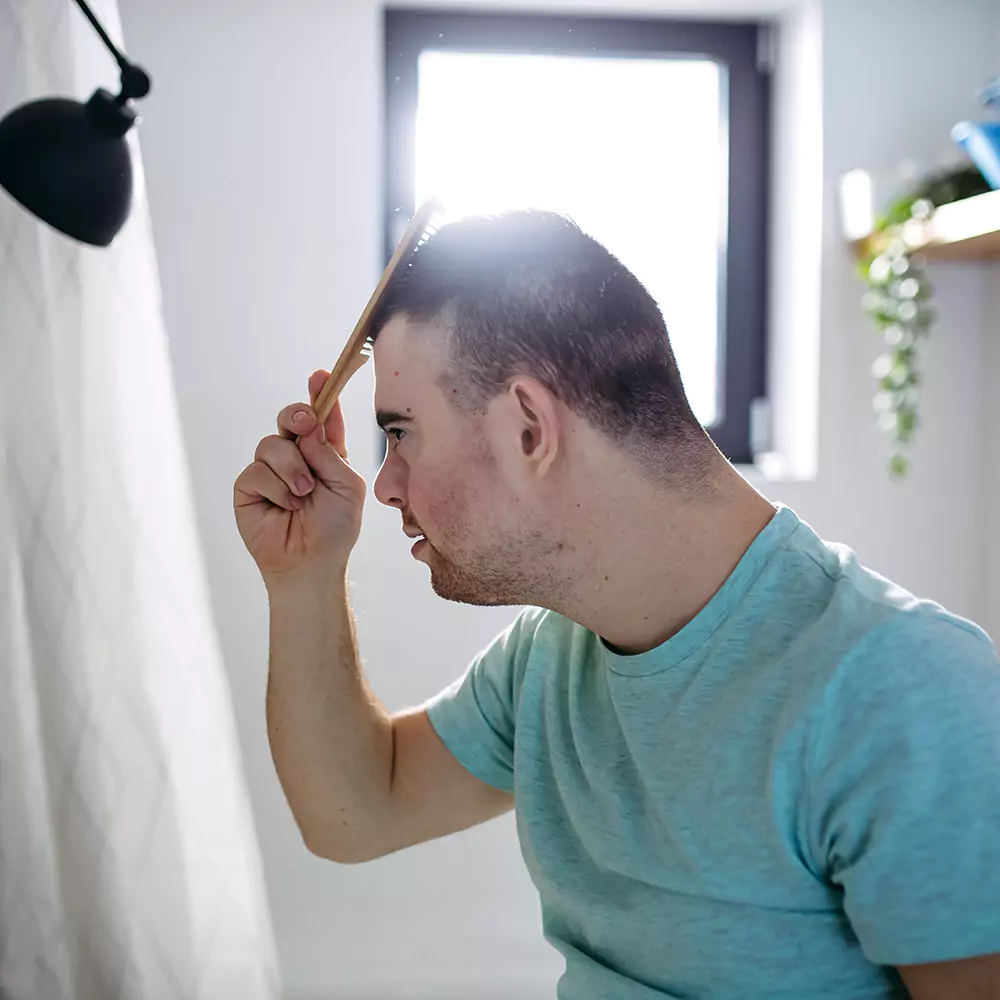 A down syndrome man with blue shirt brushing his hair in the bathroom